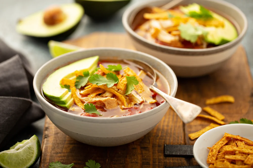 A steaming bowl of homemade taco soup garnished with shredded cheese, avocado slices, and fresh cilantro, served on a rustic wooden table.